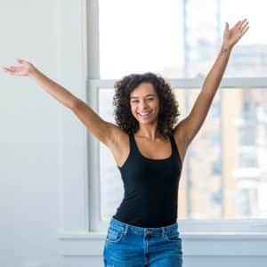 hispanic female stands in front of window of her loft or office smiling into the camera with her arms raised above her head in excitement.